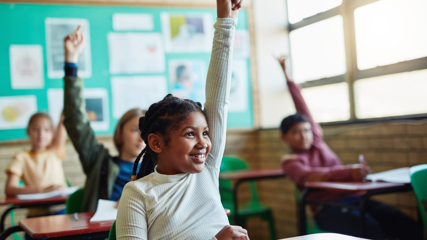 Child raising their hand sitting at a desk in a classroom